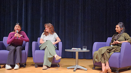 Three people are seated on a stage in purple armchairs, participating in a panel discussion. The person on the left holds a microphone and is speaking. The person in the middle listens attentively, and the person on the right, also holding a microphone, is smiling. An American flag is seen in the background.