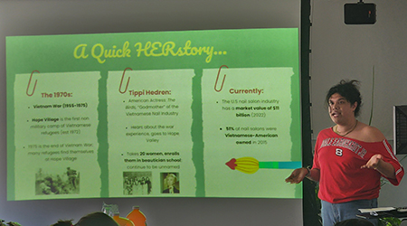 Person in a red top presenting a slideshow titled "A Quick HERstory..." to an audience. The slides discuss the nail salon industry's history and current statistics.