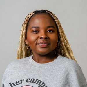 A person with blond braids and hoop earrings is wearing a gray sweatshirt with text on it. They are facing forward against a plain background.